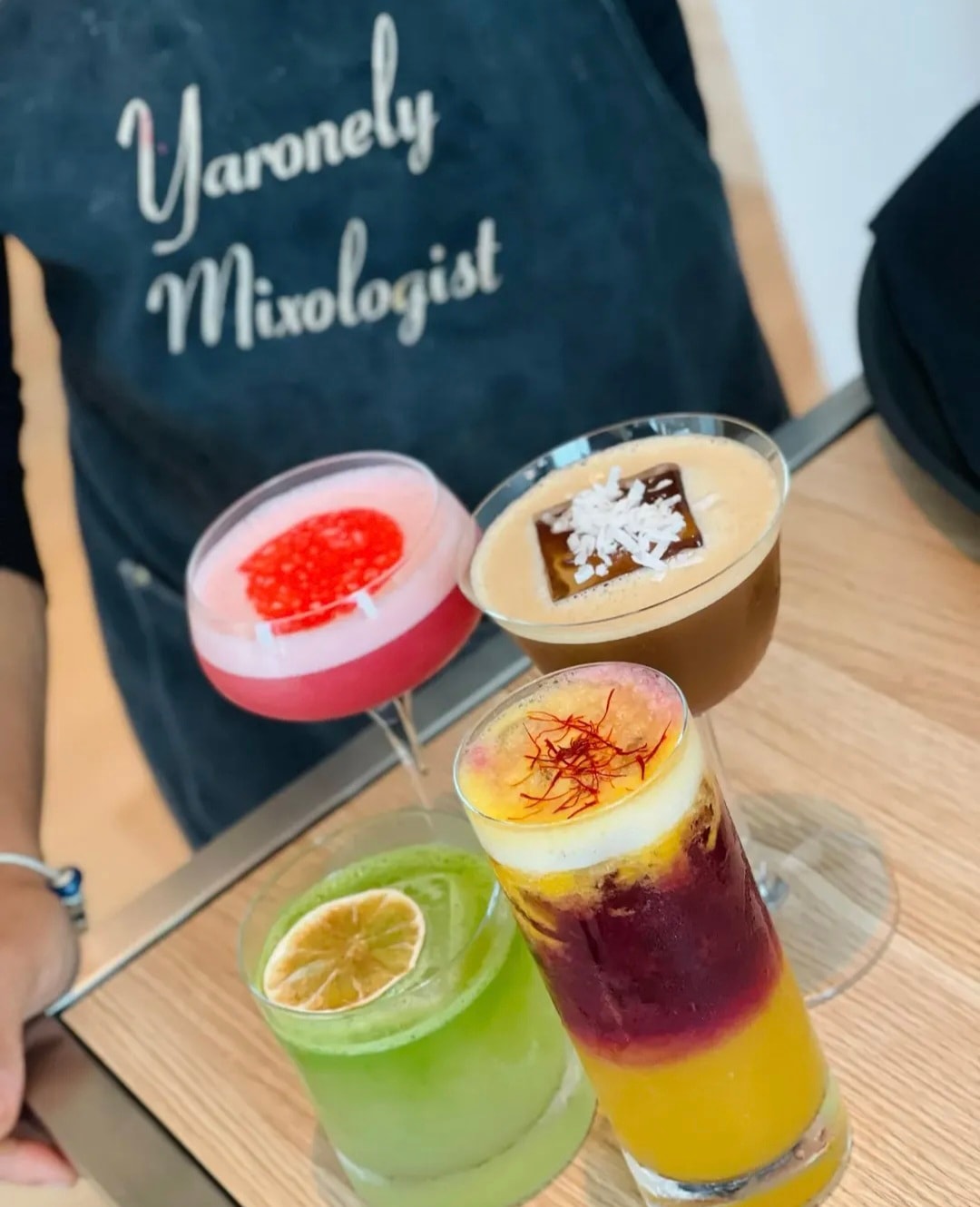Four colorful cocktails on a tray with Yaronely Mixologist apron in background